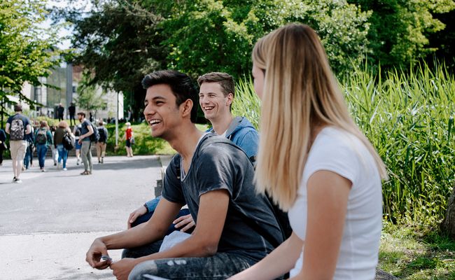 lachende Studierende, die auf Parkbank sitzen, im Hintergrund Menschen
